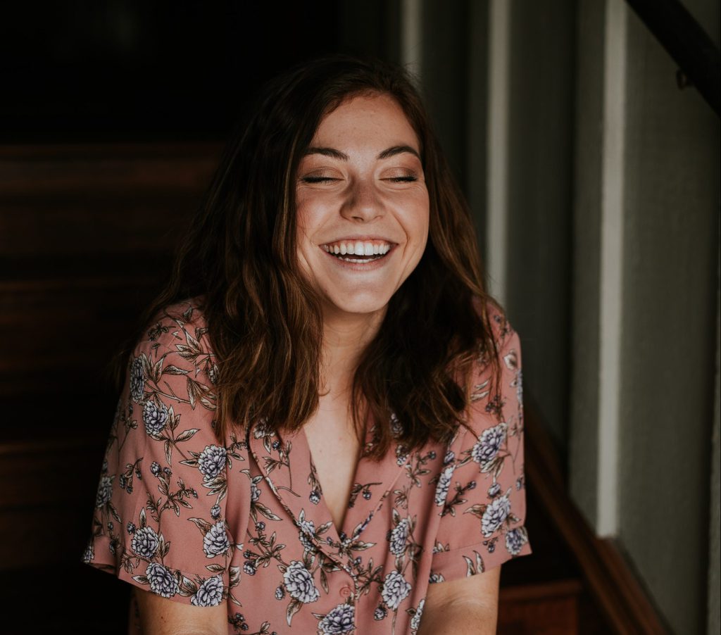 Image of: A happy and smiling young blind woman wearing a pink blouse with black and white flowers. The background is a wall that is lit by a wondow.