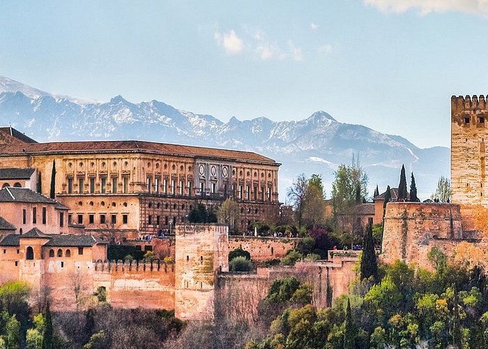 A picture showing the Alhambra, a trademark of Granada. Specifically the brown walls of a castle on the top of a hill is shown with several trees in between the openings of the walls.