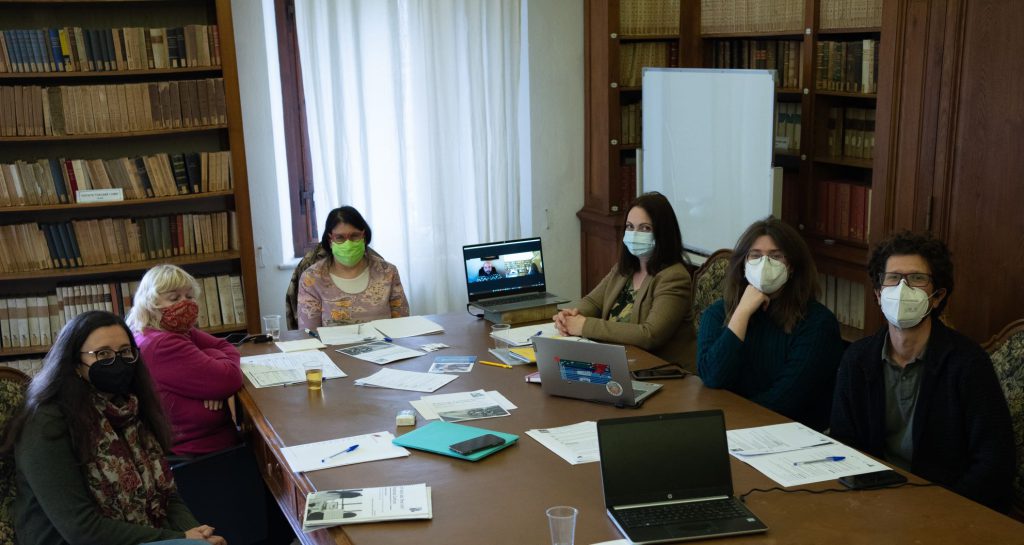 A group of 6 siting around a table with pieces of paper scattered on it. In the table there is also a computer that is open and shows that one person is joining the meeting online. The room is a library and the walls are full of books. There is a window in the middle of the background wall.