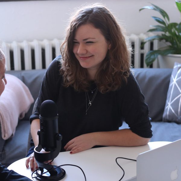 two women siting behind a table that on it has a microphone and a laptop. We cans ee the back of one and the face of the other. The face that is shown is smiling.